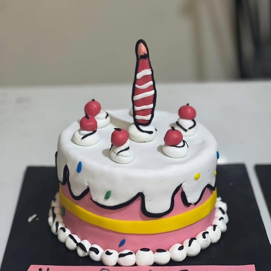 A round pink and white cake with dark outlines, red toppers, and a tall striped candle.