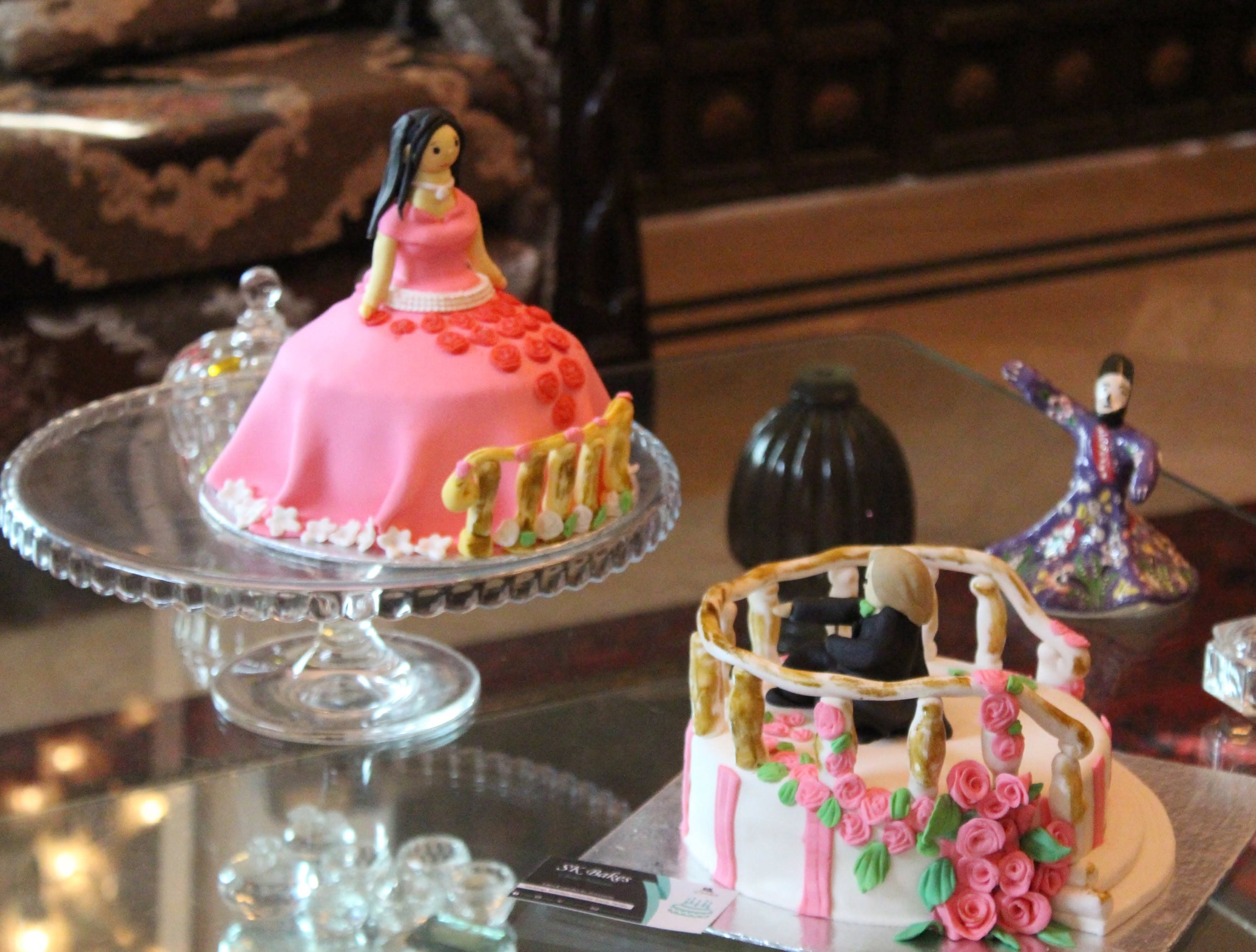 A pink doll-shaped cake displayed alongside another pink decorated celebration cake. Alternate view.
