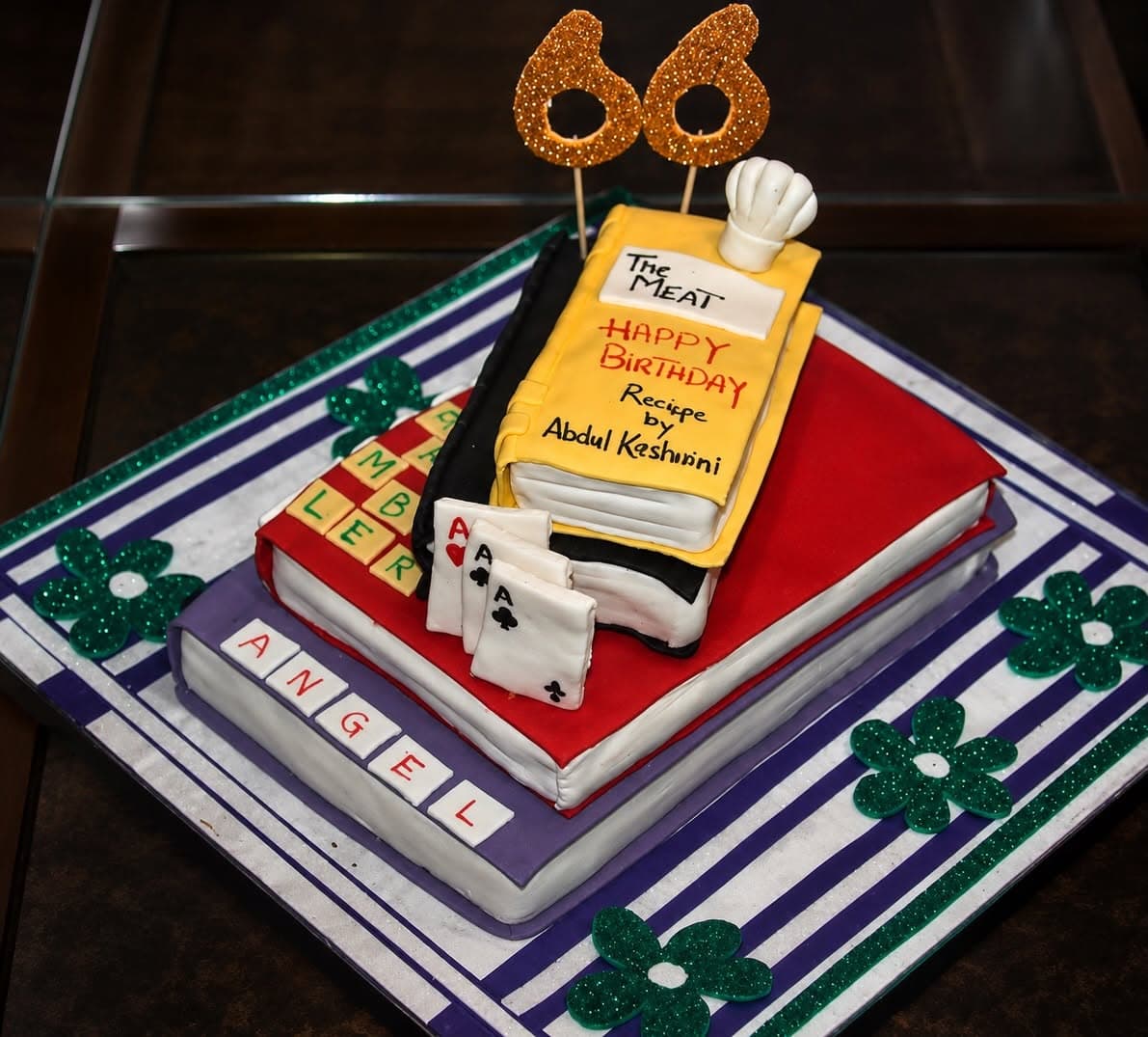 A custom cake shaped as stacked books, with gold number toppers and miniature decorative details. Alternate view.